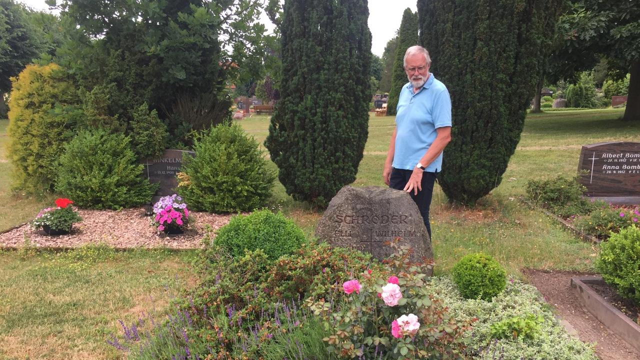 Ein grauhaariger Mann mit hellblauem, kurzärmeligen Hemd steht auf einem Friedhof vor einem Grab, das von üppig gewachsenen Pflanzen bedeckt ist. Ein grauhaariger Mann mit hellblauem, kurzärmeligen Hemd steht auf einem Friedhof vor einem Grab, das von üppig gewachsenen Pflanzen bedeckt ist.