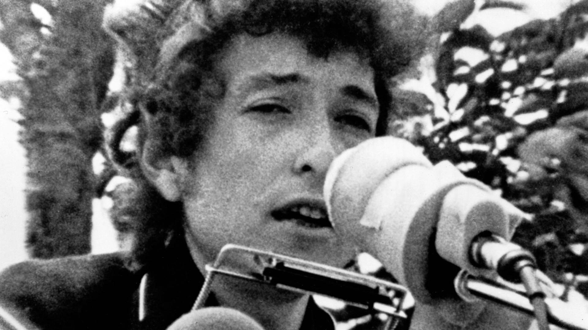 Bob Dylan auf einem grobkörnigen Schwarz-Weiß-Foto aus den 1960er Jahren. Er hat eine Mundharmonika und ein Mikro vorm Gesicht.