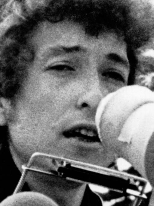 Bob Dylan auf einem grobkörnigen Schwarz-Weiß-Foto aus den 1960er Jahren. Er hat eine Mundharmonika und ein Mikro vorm Gesicht.