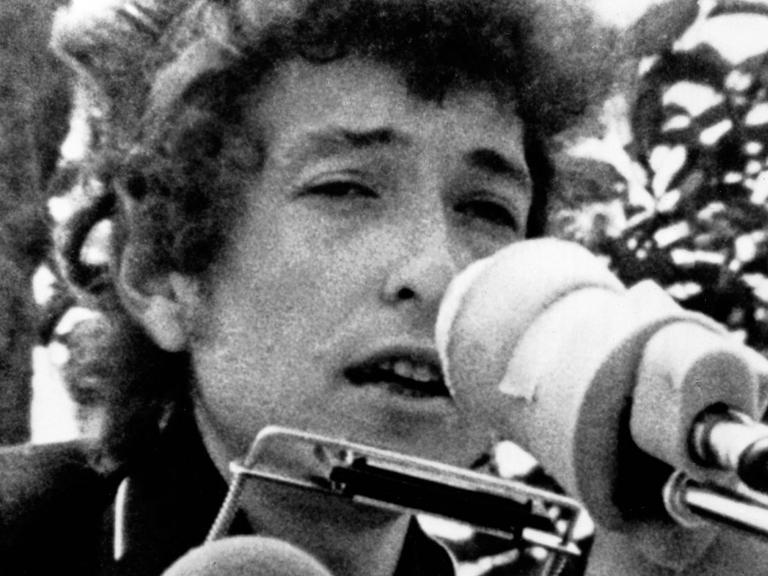 Bob Dylan auf einem grobkörnigen Schwarz-Weiß-Foto aus den 1960er Jahren. Er hat eine Mundharmonika und ein Mikro vorm Gesicht.
