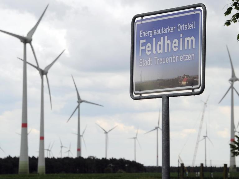 Ein blaues Schild, hinter dem sich Windräder drehen, weist Feldheim (Brandenburg) am 08.05.2014 als energieautarken Ort aus.