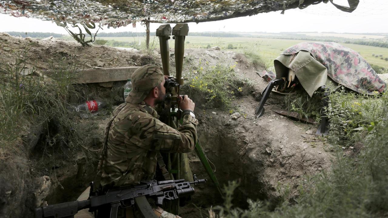 Der Soldat sitzt in einem von einem Tarnzelt überdachten Schützengraben und schaut durch ein Fernsichtgerät. 