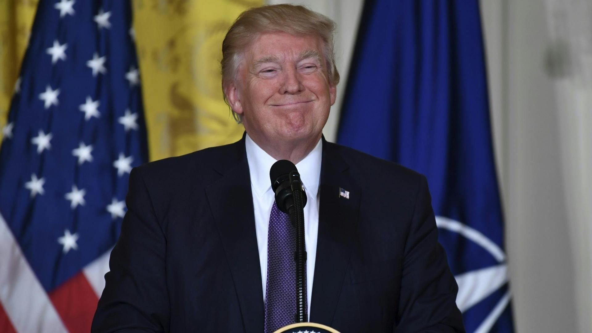 US-Präsident Trump steht an einem Rednerpult und grinst breit, hinter ihm die Flaggen der USA und der Nato. US-Präsident Trump steht an einem Rednerpult und grinst breit, hinter ihm die Flaggen der USA und der Nato.
