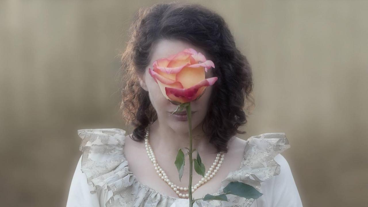 Eine Frau hält sich eine große gelb-rote Rose vor das Gesicht. Sie trägt ein historisches Kleid, eine Perlenkette und weiße Handschuhe. Eine Frau hält sich eine große gelb-rote Rose vor das Gesicht. Sie trägt ein historisches Kleid, eine Perlenkette und weiße Handschuhe.