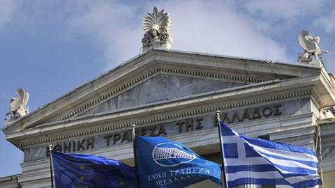 Vor dem Gebäude der griechischen Nationalbank in Athen flattern Flaggen der EU, der Bank und Griechenlands im Wind.