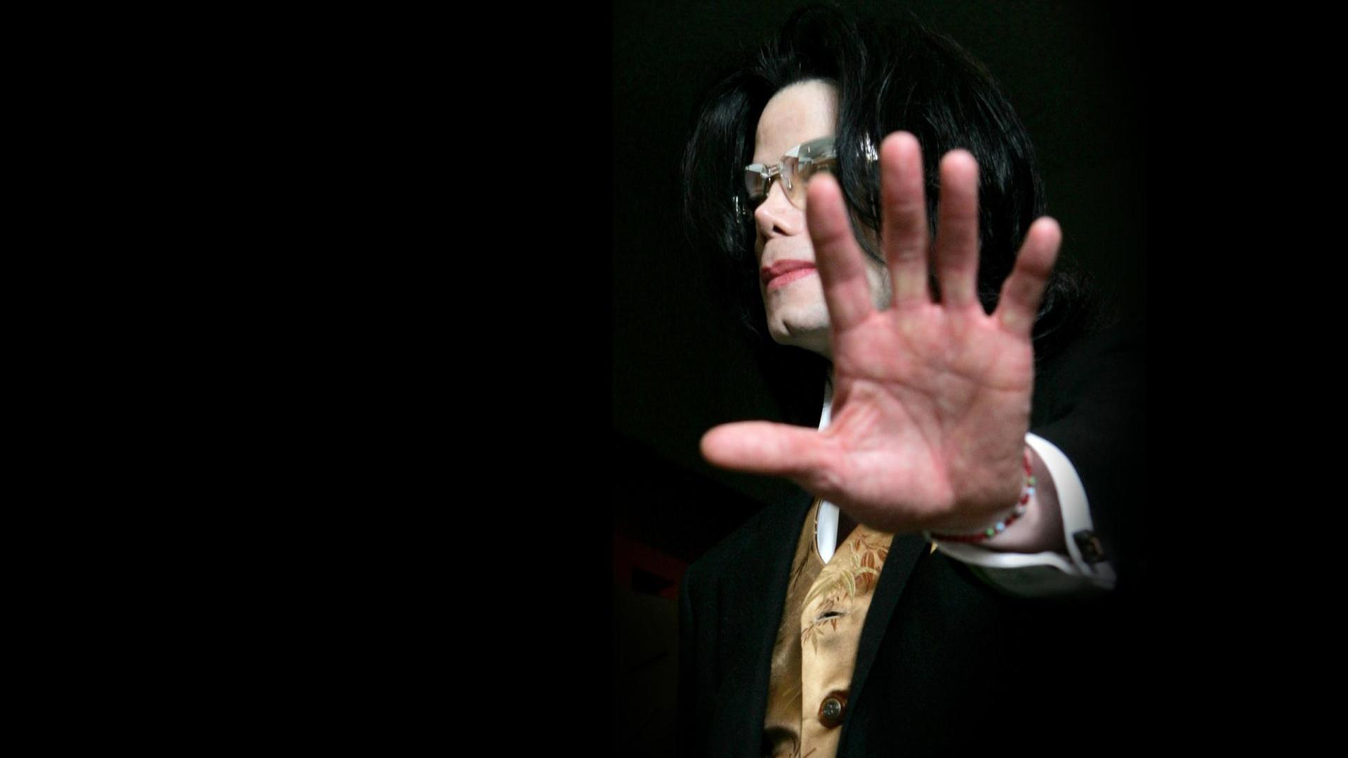 Michael Jackson streckt die Handlfäche der linken Hand Richtung Kamera. Er trägt eine Sonnenbrille. Der Hintergrund ist schwarz.