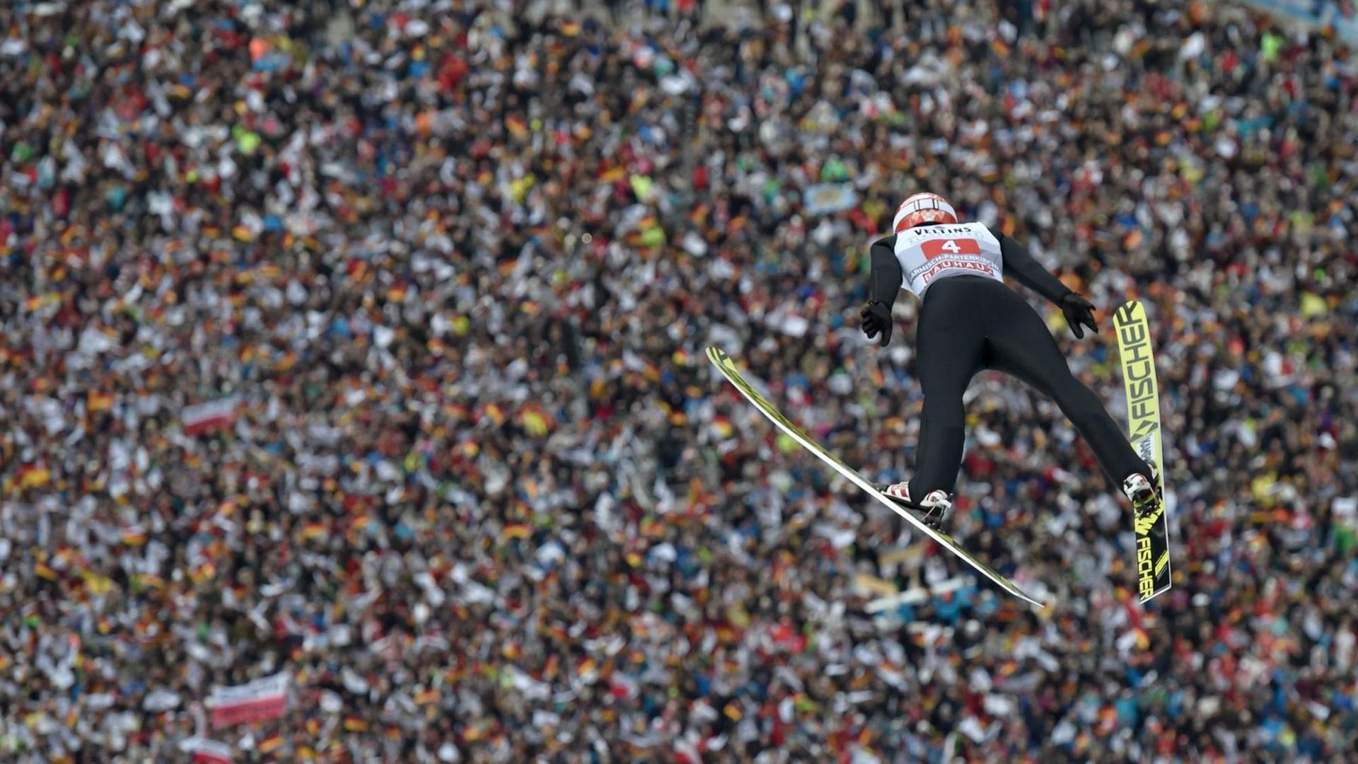 Das Bild zeigt Skispringe Markus Eisenbichler nach dem Sprung von der Schanze in der Luft. Im Hintergrund ist eine Zuschauer-Menge zu sehen. Das Bild zeigt Skispringe Markus Eisenbichler nach dem Sprung von der Schanze in der Luft. Im Hintergrund ist eine Zuschauer-Menge zu sehen.