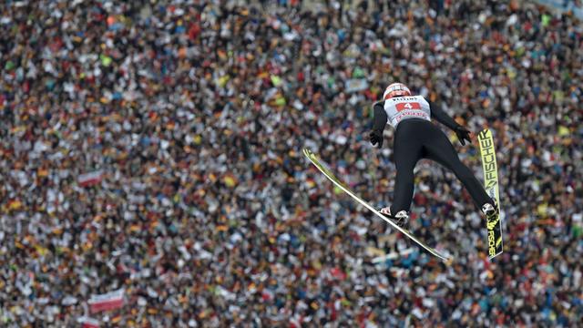 Das Bild zeigt Skispringe Markus Eisenbichler nach dem Sprung von der Schanze in der Luft. Im Hintergrund ist eine Zuschauer-Menge zu sehen.