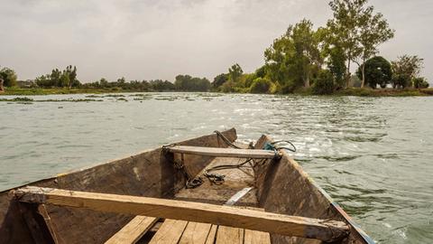 Im Hinterland, wo einmal die Geheime Stadt gestanden hat, wohnt Bowguard. Zu sehen: Die Spitze eines alten Holzboots auf dem Niger. Am Ufer sind Bäume und eine Graslandschaft.