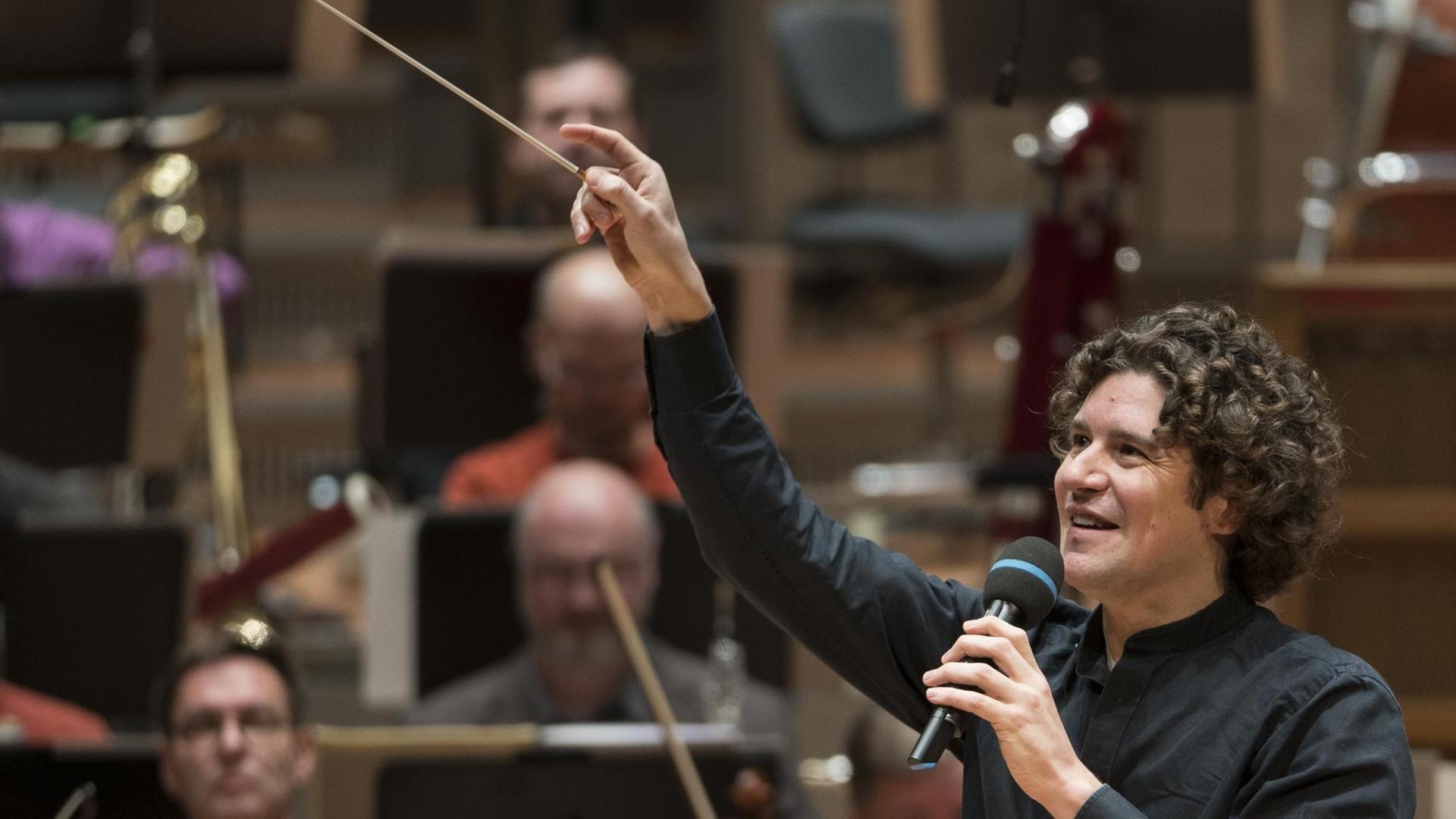 Dirigent Robin Ticciati mit Taktstock und Mikrofon in der Hand moderiert ein Konzert in der Berlliner Philharmonie. Dirigent Robin Ticciati mit Taktstock und Mikrofon in der Hand moderiert ein Konzert in der Berlliner Philharmonie.