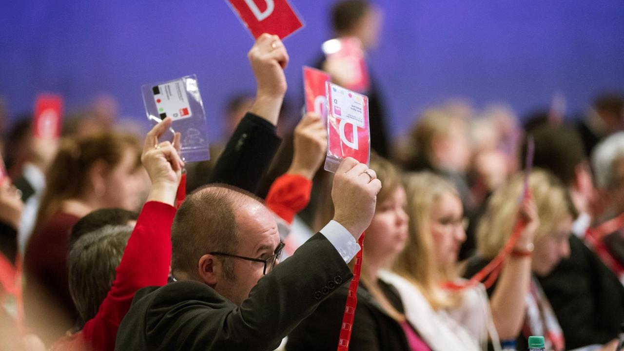 Das Bild zeigt Delegierte von der Seite, die an ihren Tischen sitzen und rote Stimmkarten nach oben halten.