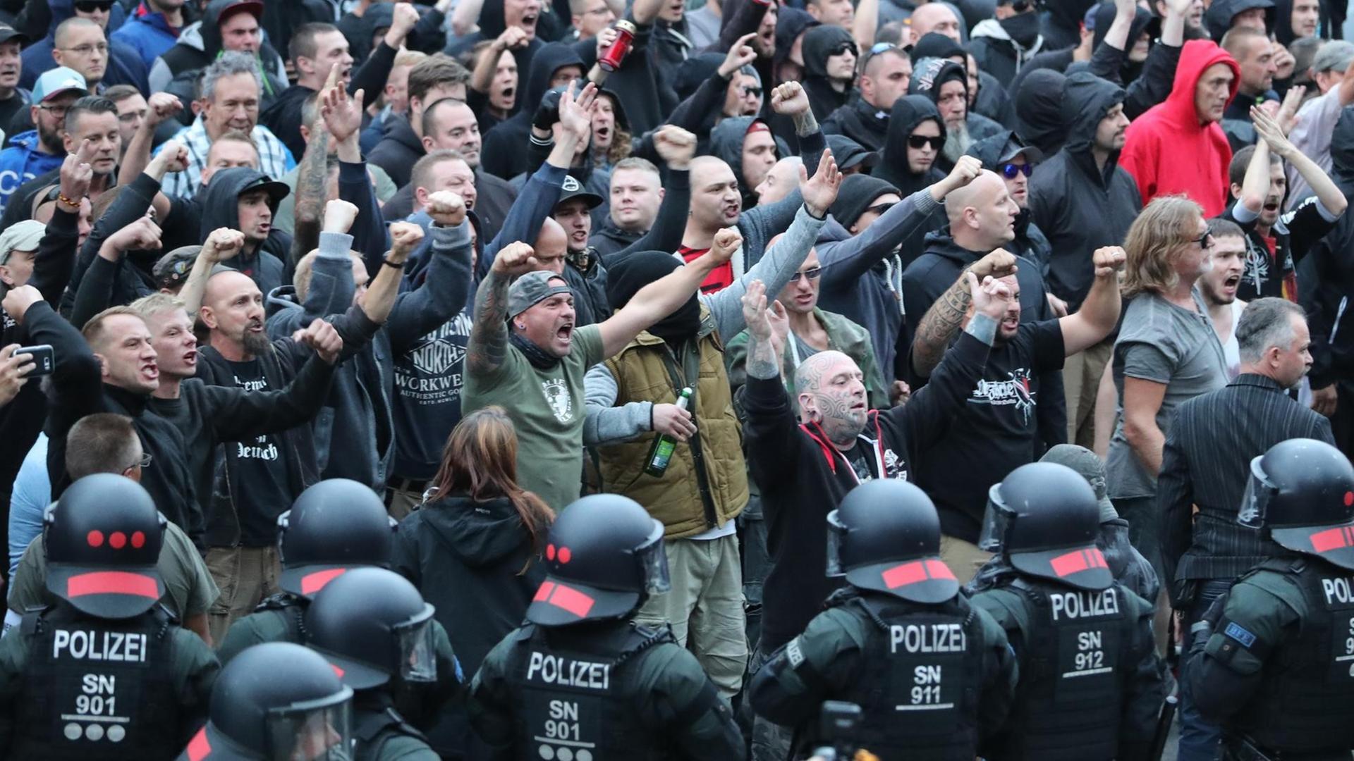 Das Bild zeigt am unteren Rand schwarzgekleidete Polizisten in Kampfmontur mit Helm. Hinter ihnen eine große Menge rechtsgerichteter Demonstranten, viele rufen etwas und halten die geballten Fäuste nach oben. Man sieht viele hassverzerrte Gesichter unter ihnen.