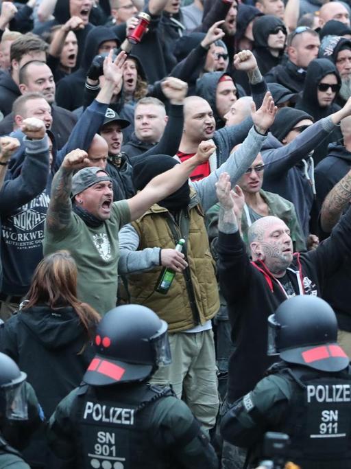 Das Bild zeigt am unteren Rand schwarzgekleidete Polizisten in Kampfmontur mit Helm. Hinter ihnen eine große Menge rechtsgerichteter Demonstranten, viele rufen etwas und halten die geballten Fäuste nach oben. Man sieht viele hassverzerrte Gesichter unter ihnen.