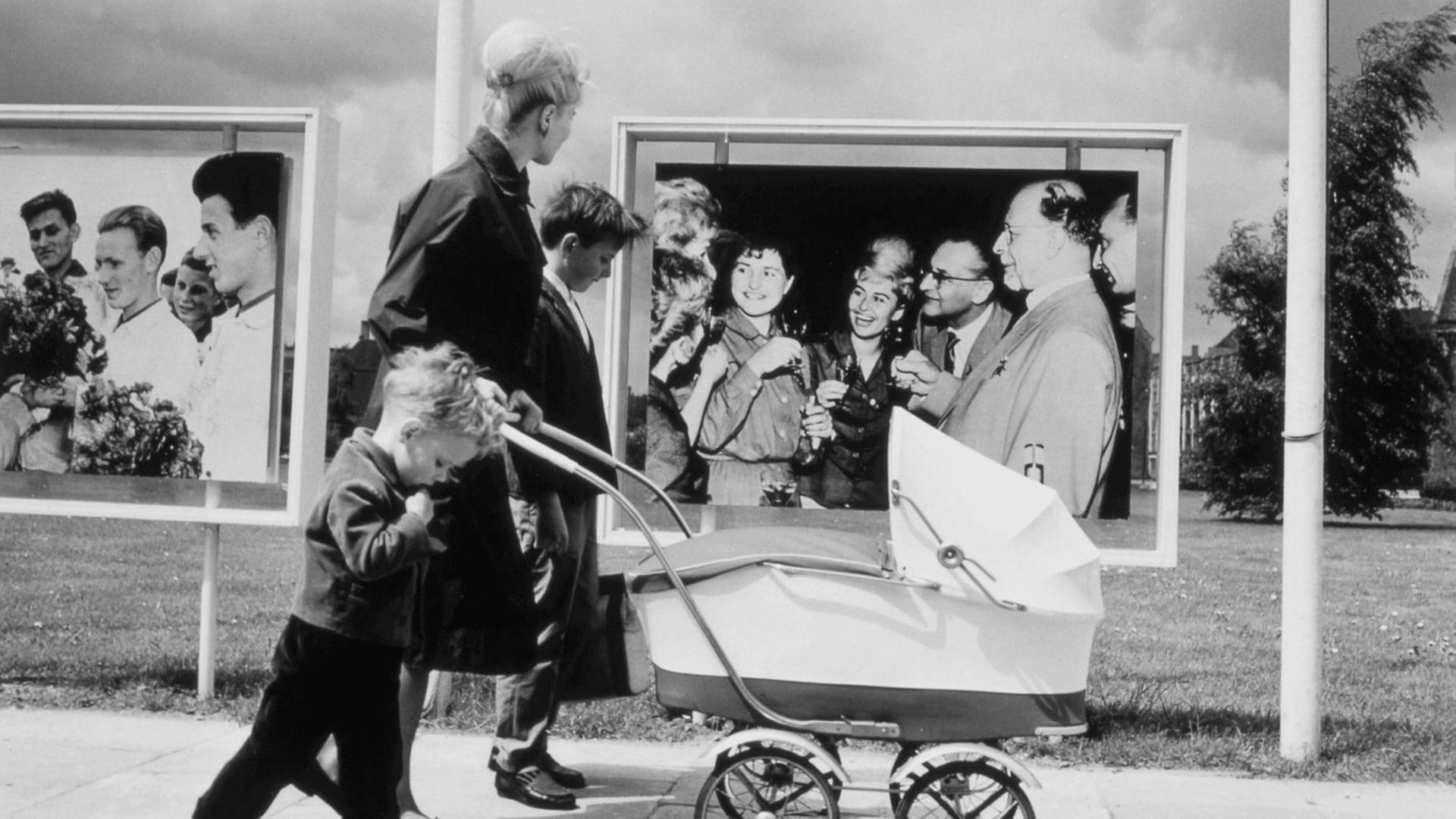 Mutter mit ihren Kindern und einem Kinderwagen, unterwegs in Ostberlin, vor Fotowänden mit SED Propaganda (Fotowand rechts: SED-Generalsekretär Walter Ulbricht u. a.). Foto, 1964. | Mutter mit ihren Kindern und einem Kinderwagen, unterwegs in Ostberlin, vor Fotowänden mit SED Propaganda (Fotowand rechts: SED-Generalsekretär Walter Ulbricht u. a.). Foto, 1964. |