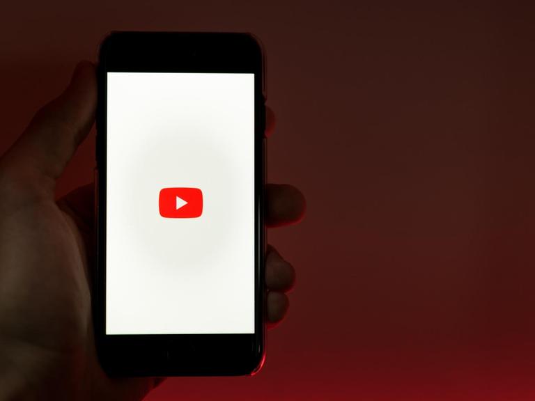 Eine Hand hält ein Smartphone in die Kamera, auf dem das Logo von Youtube abgebildet ist.
