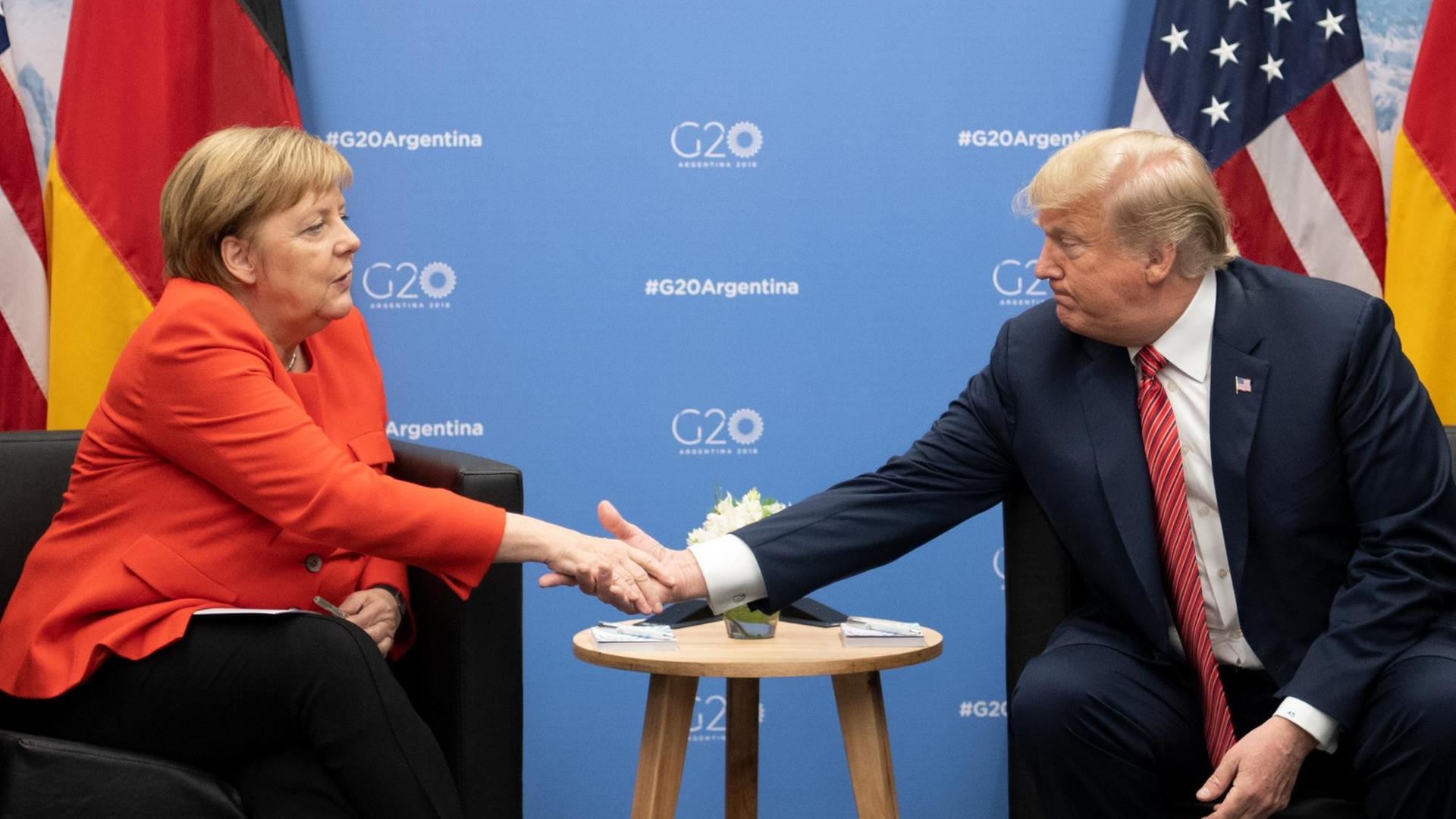 Das Bild zeigt Bundeskanzlerin Angela Merkel (CDU) und Donald Trump, Präsident der Vereinigten Staaten, Sie sitzen nebeneinander an einem Tisch und geben sich die Hand. Im Hinergrund sind die Flaggen Deutschlands und der USA sowie eine blaue Wand mit dem offiziellen Logo des Gipfeltreffens zu sehen. Das Bild zeigt Bundeskanzlerin Angela Merkel (CDU) und Donald Trump, Präsident der Vereinigten Staaten, Sie sitzen nebeneinander an einem Tisch und geben sich die Hand. Im Hinergrund sind die Flaggen Deutschlands und der USA sowie eine blaue Wand mit dem offiziellen Logo des Gipfeltreffens zu sehen.