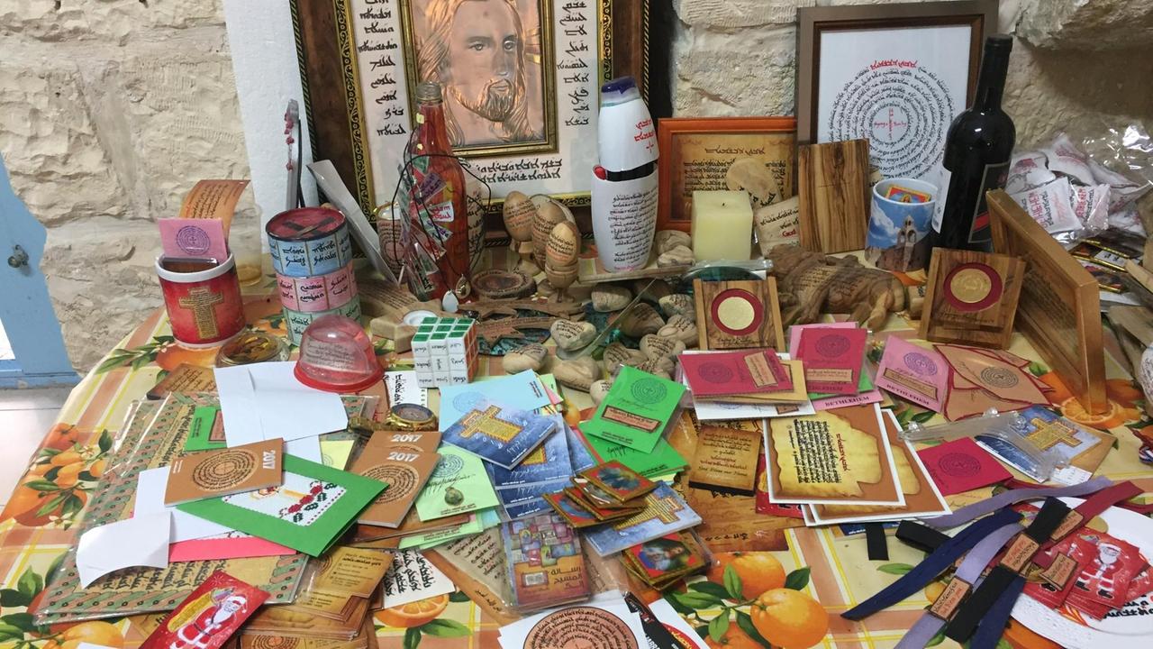 Auf einem Tisch liegen durcheinander viele verschiedene Geschenkartikel mit aramäischen Schriften: Herzen aus Olivenholz mit dem Vaterunser auf aramäisch, Sticker, Kühlschrankmagneten, Tassen, Kalender, Weihnachtskarten, Bilder, ein Zauberwüfel.