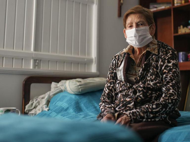 Eine ältere Dame mit Mundschutz sitzt alleine auf einem Bett. Auch in Bogota, Kolumbien werden alle älteren Menschen ab 70 Jahren gebeten, ihr Haus nicht zu verlassen. 18. März 2020