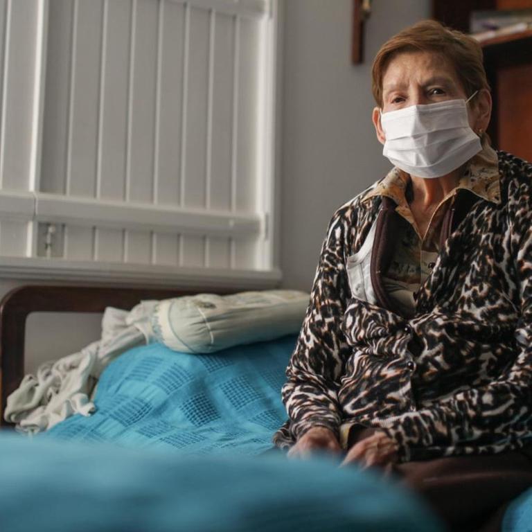 Eine ältere Dame mit Mundschutz sitzt alleine auf einem Bett. Auch in Bogota, Kolumbien werden alle älteren Menschen ab 70 Jahren gebeten, ihr Haus nicht zu verlassen. 18. März 2020