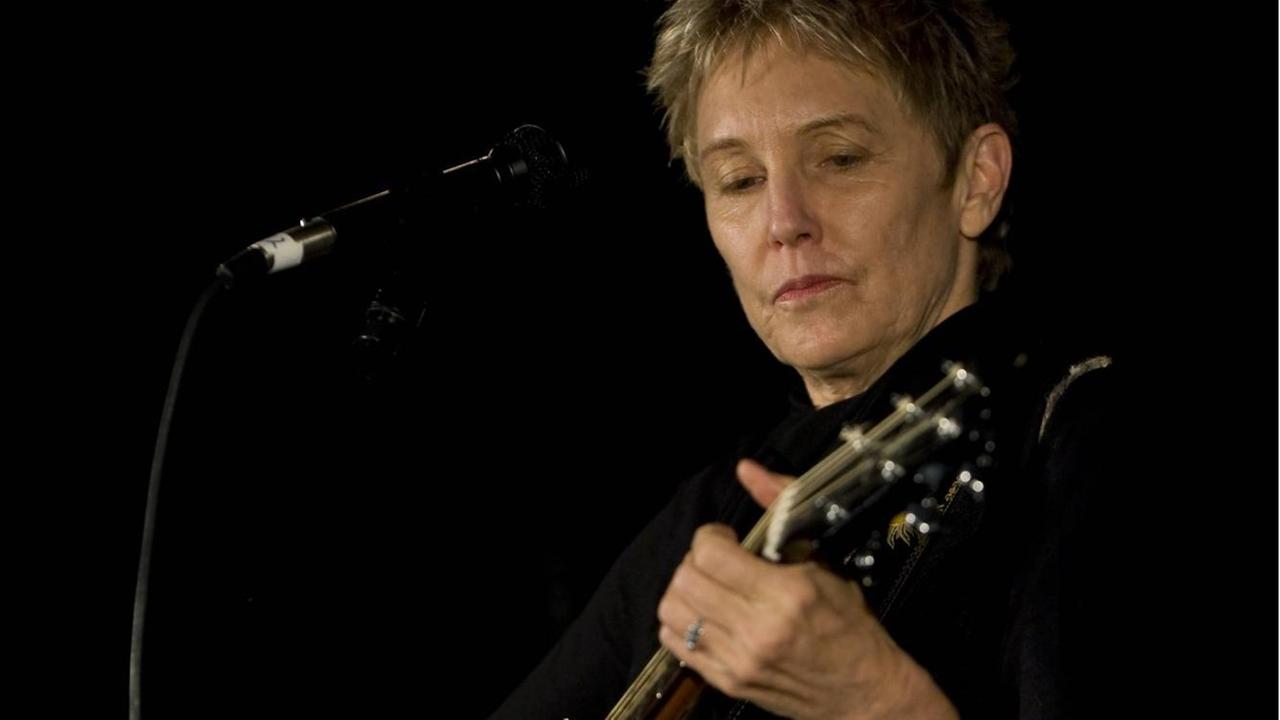Eine Frau mit kurzen grauen Haaren steht auf einer Bühne mit Mikrofon und spielt Gitarre.
