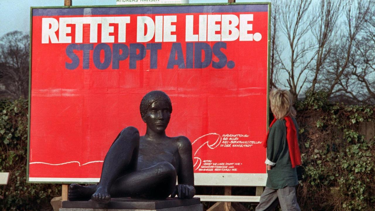Ein Frau geht an einem roten Plakat mit dem Spruch "Rettet die Liebe - Stoppt Aids" vorbei. Im Vordegrund ist eine liegende Frauenskulptur zu sehen.