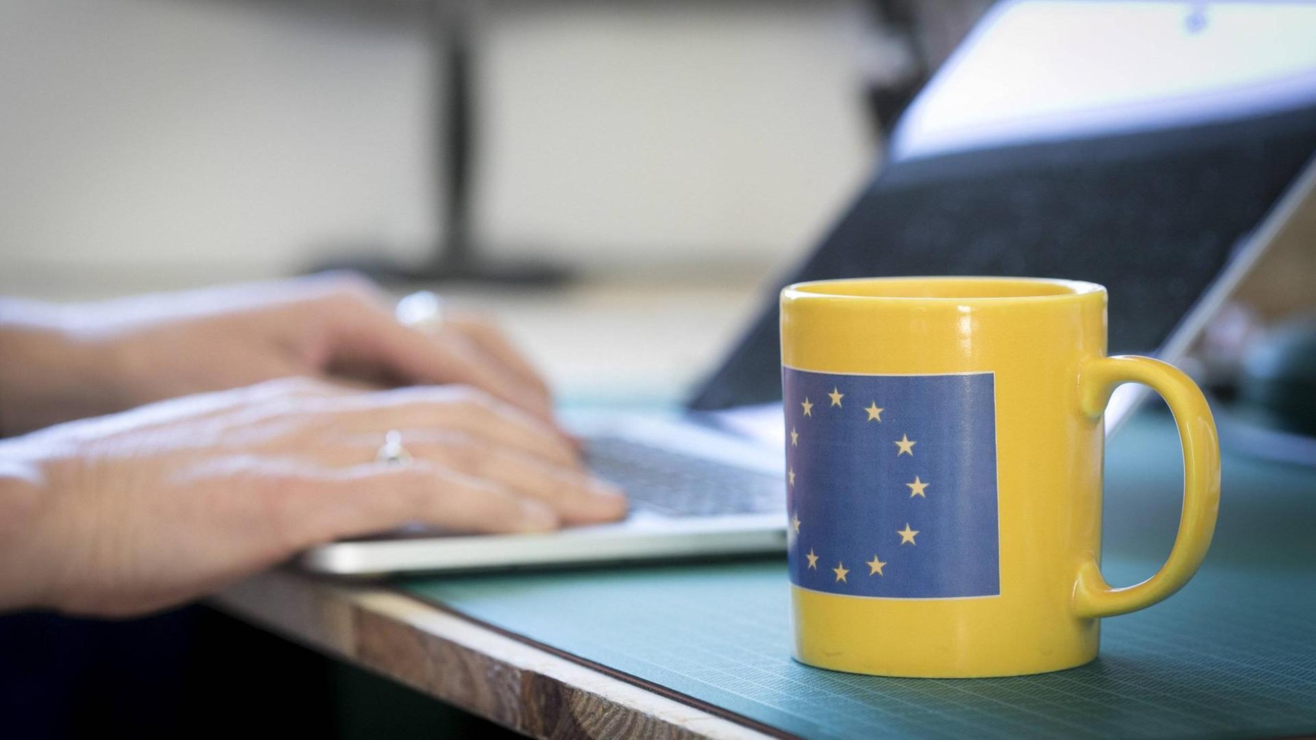 Eine Tasse mit der Flagge Europas Schreibtisch, an dem eine Person am Laptop sitzt. Eine Tasse mit der Flagge Europas Schreibtisch, an dem eine Person am Laptop sitzt.