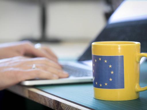 Eine Tasse mit der Flagge Europas Schreibtisch, an dem eine Person am Laptop sitzt.