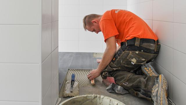 Ein Mann im orangefarbenen T-Shirt verlegt große graue Fußbodenfliesen in einem Bad.