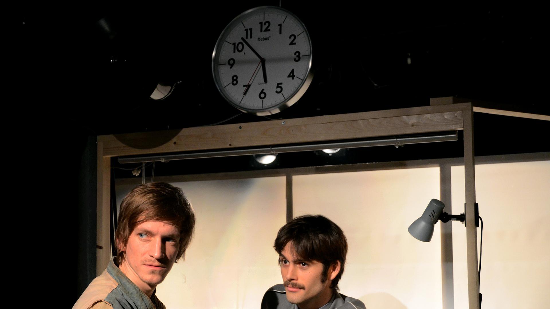 Schauspieler Lasse Marburg lehnt an einem Kontrollpunkt, hinter dem Schauspieler Philip Steimel steht, beide schauen links an der Kamera vorbei. Über ihnen zeigt eine analoge Uhr 5.52 Uhr an.