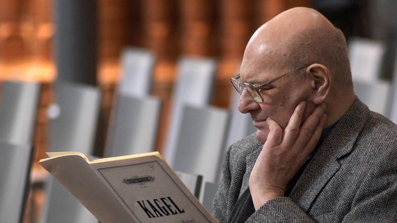 Der kahlköpfige Komponist sitzt mit einem Notenheft, auf dem mit großen Buchstaben "Kagel" steht, in den leeren Reihen eines modernen, hellen Zuschauerraumes. Der kahlköpfige Komponist sitzt mit einem Notenheft, auf dem mit großen Buchstaben "Kagel" steht, in den leeren Reihen eines modernen, hellen Zuschauerraumes.