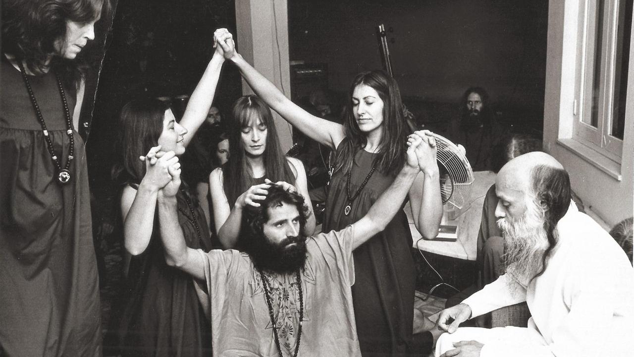 Ein Mann mit langen Haaren und Vollbart schaut ehrfürchtig den indischen Guru Bhagwan an, der rechts von ihm in einem weißen Gewand sitzt, drei Frauen erheben in einer rituellen Geste ihre Arme über dem Mann, eine legt ihm ihre Hände auf den Kopf.