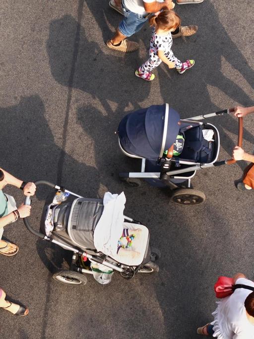 Vogelperspektive: Zwei junge Familien mit Kindern begegenen sich, als sie über eine Straße gehen. Bei der einen Familie schiebt die Mutter den Kinderwagen, bei der anderen der Vater.