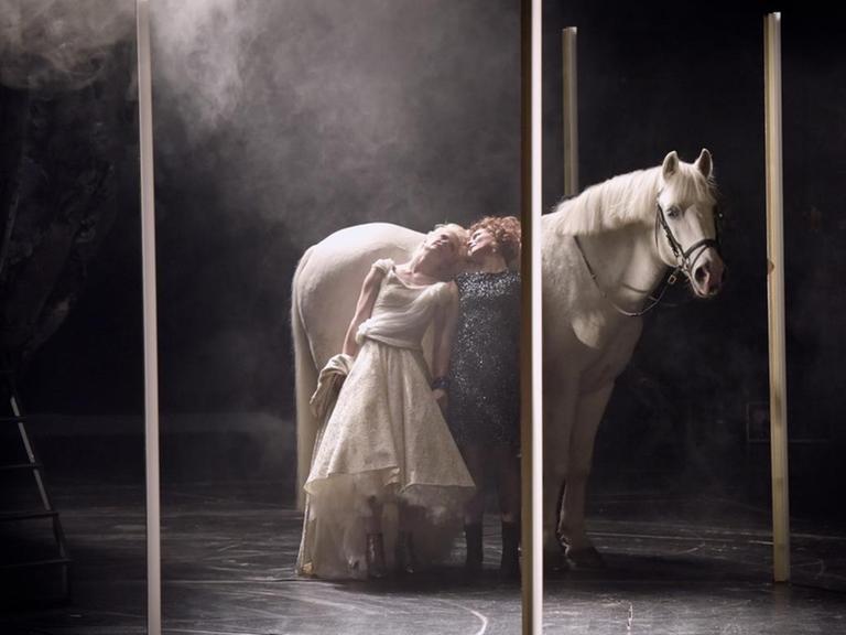 Eine Szene aus "Melancholia" nach dem Film von Lars von Trier am Schauspielhaus Bochum: Ein weisses Pferd und zwei Schauspielerinnen stehen zwischen Stelen auf der Bühne.