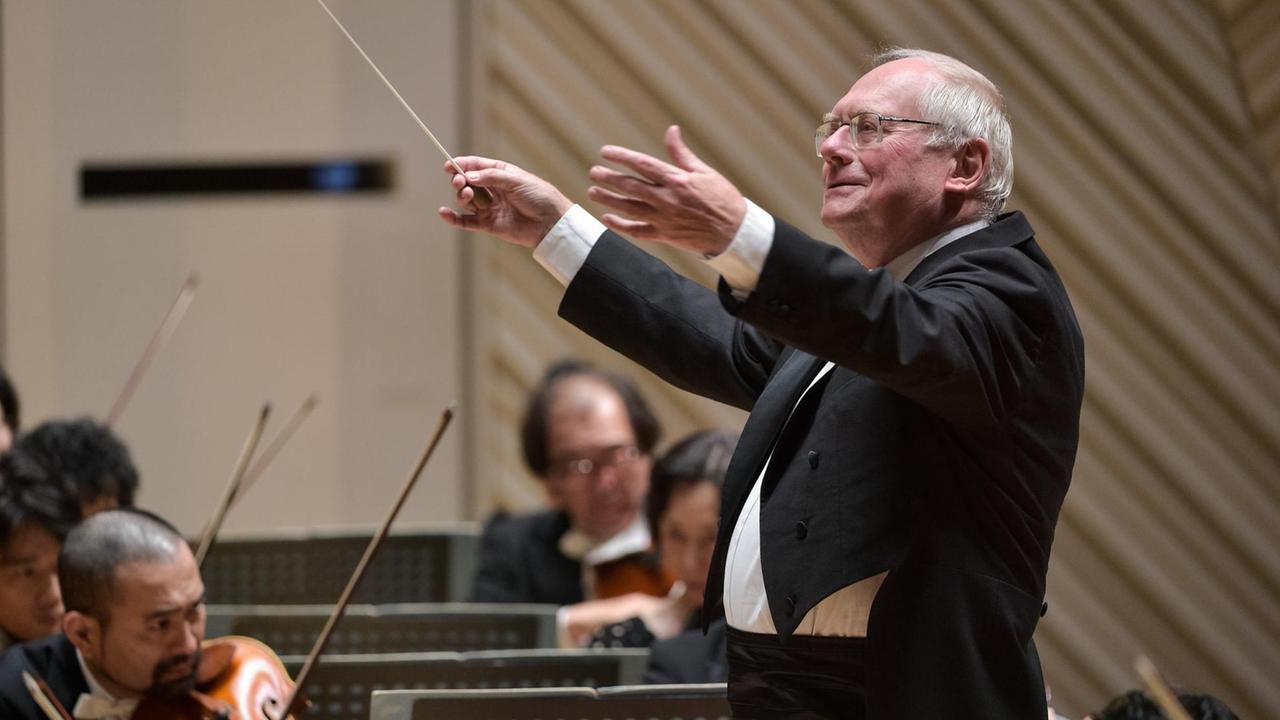 Ein älterer Mann mit Brille und weißem Haar dirigiert ein Orchester und lächelt dabei.