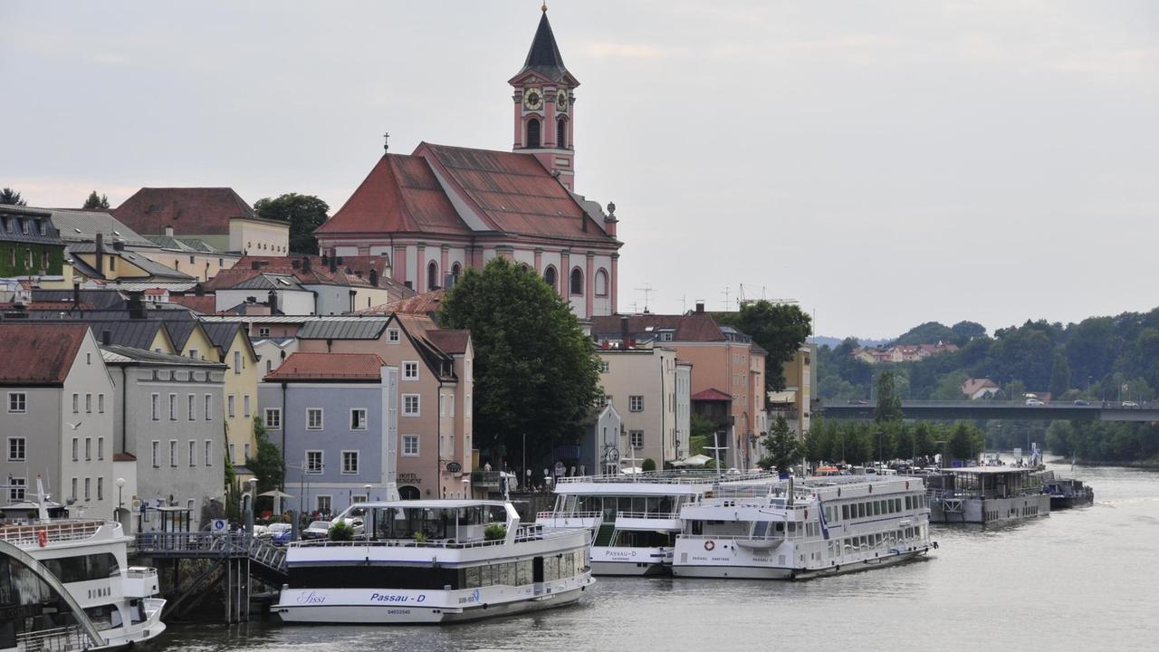 Blick auf die Schiffsanleger und Pfarrkirche St. Paul in Passau. Mehrere Kreuzfahrtschiffe stehen dicht gedrängt am Donauufer.