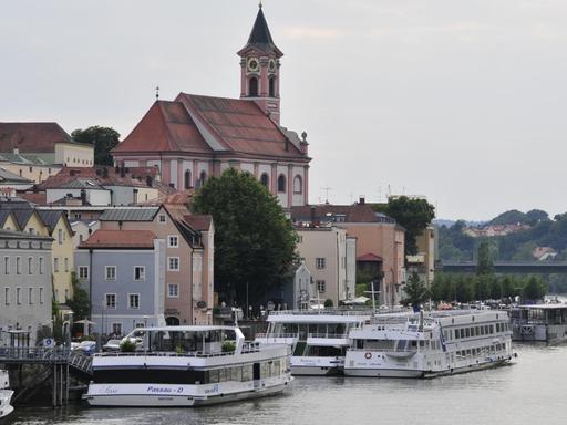 Blick auf die Schiffsanleger und Pfarrkirche St. Paul in Passau. Mehrere Kreuzfahrtschiffe stehen dicht gedrängt am Donauufer.