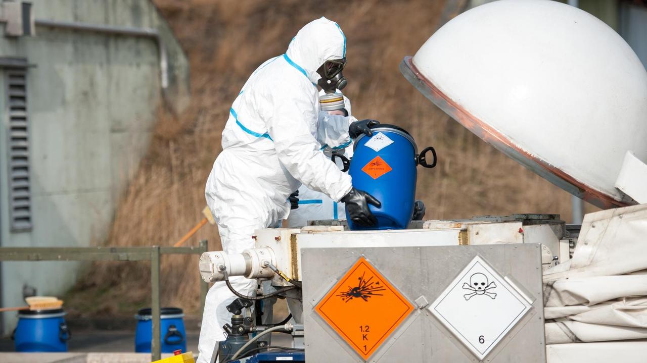 Der Mann mit weißem Schutzanzug und Gasmaske holt einen blauen Behälter aus einer Kiste mit Kampfstoff-Warnhinweisen. Der Mann mit weißem Schutzanzug und Gasmaske holt einen blauen Behälter aus einer Kiste mit Kampfstoff-Warnhinweisen.