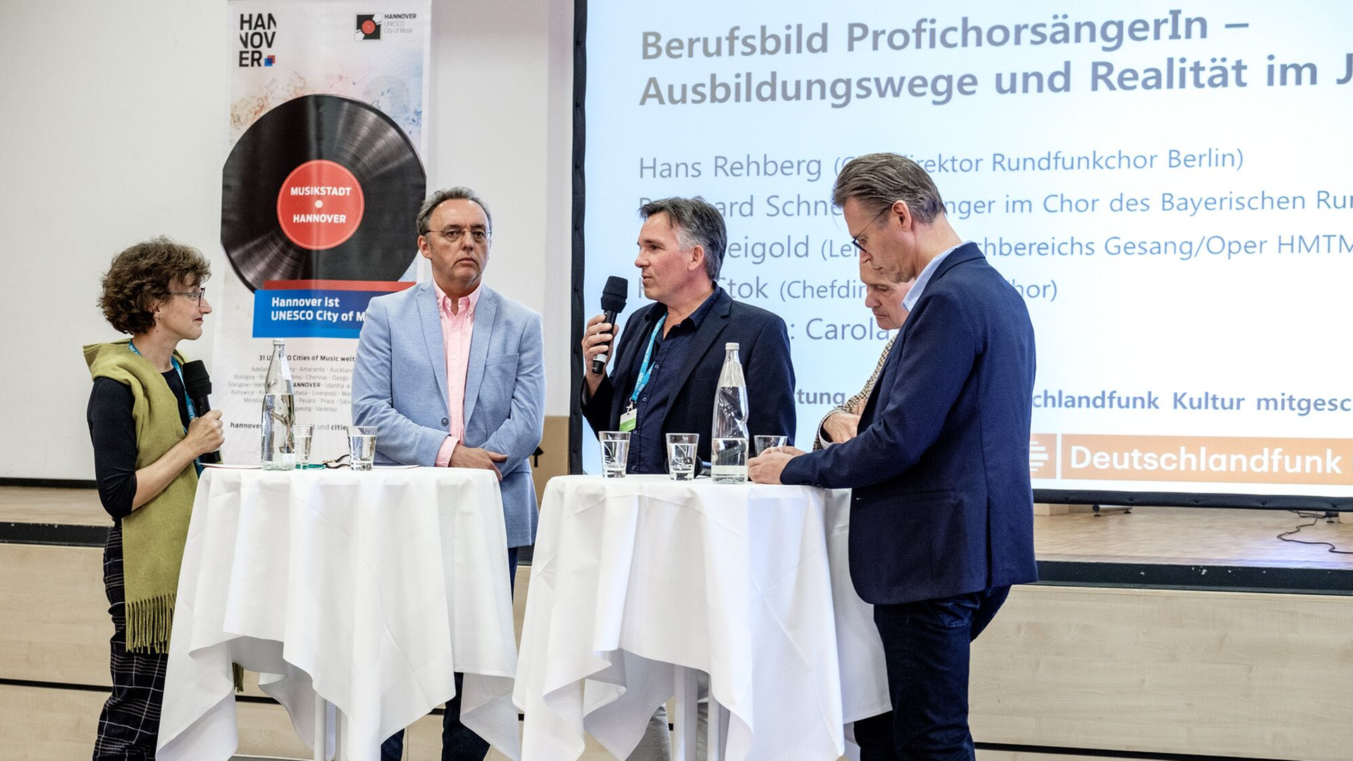Diskussionteilnehmer stehen auf einer Bühne an zwei weißen Stehtischen Diskussionteilnehmer stehen auf einer Bühne an zwei weißen Stehtischen