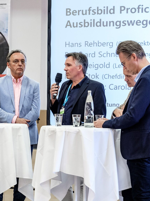 Diskussionteilnehmer stehen auf einer Bühne an zwei weißen Stehtischen
