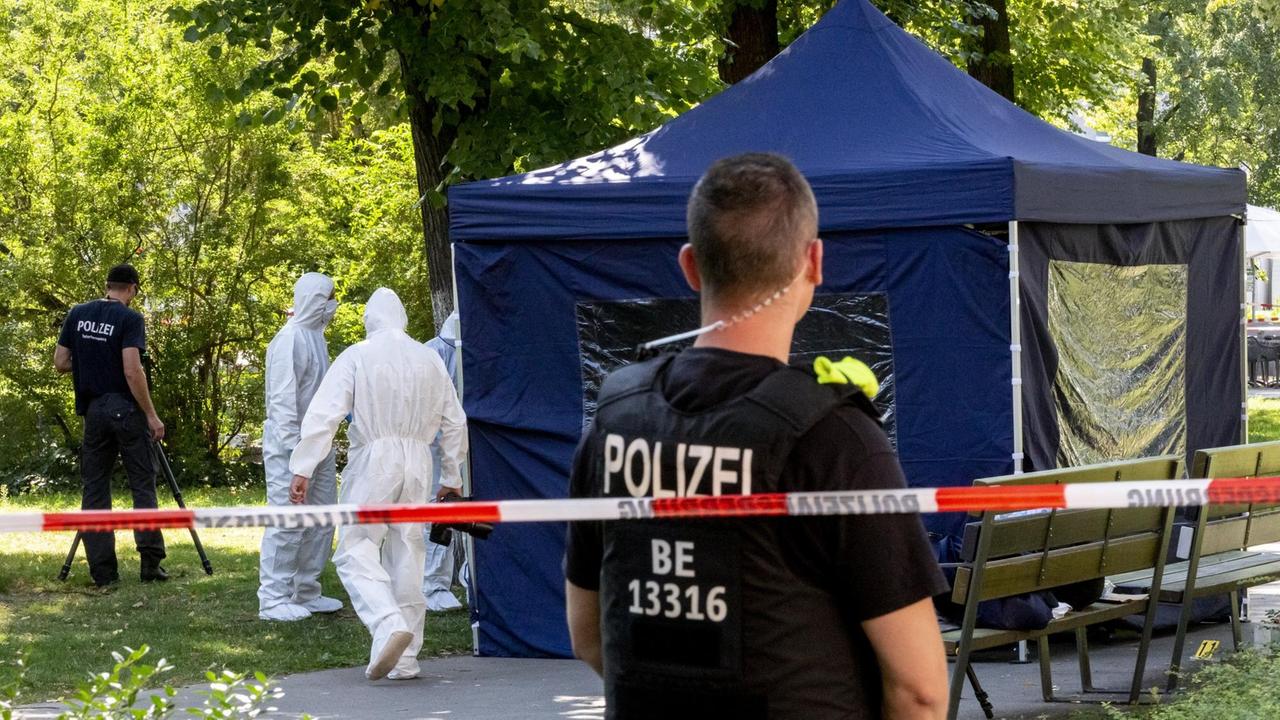 Ein Polizist steht mit dem Rücken zur Kamera hinter einem rot-weißen Absperrband. Er blickt auf ein blaues Zelt und auf Ermittler in weißen Schutzanzügen. Ein Polizist steht mit dem Rücken zur Kamera hinter einem rot-weißen Absperrband. Er blickt auf ein blaues Zelt und auf Ermittler in weißen Schutzanzügen.