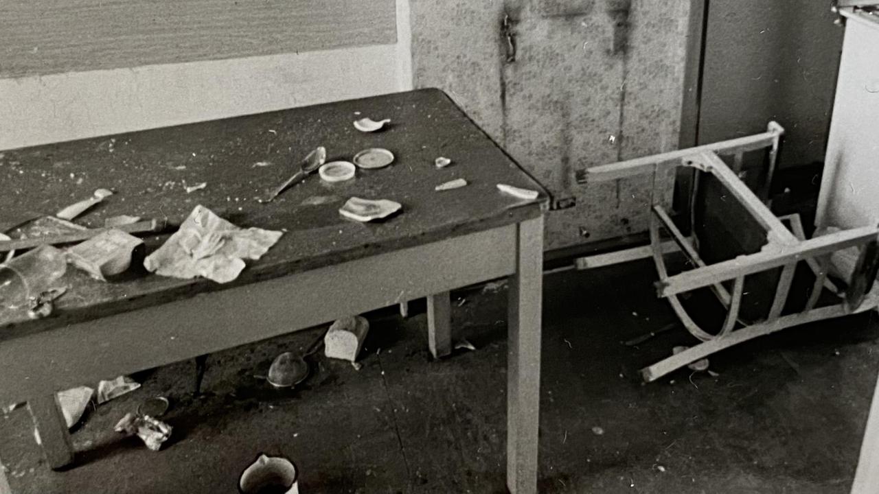 Schwarz-Weiß Aufnahme eines umgestoßenen Stuhls und eines Tisches der mit zerbrochenen Geständen überstreut ist.