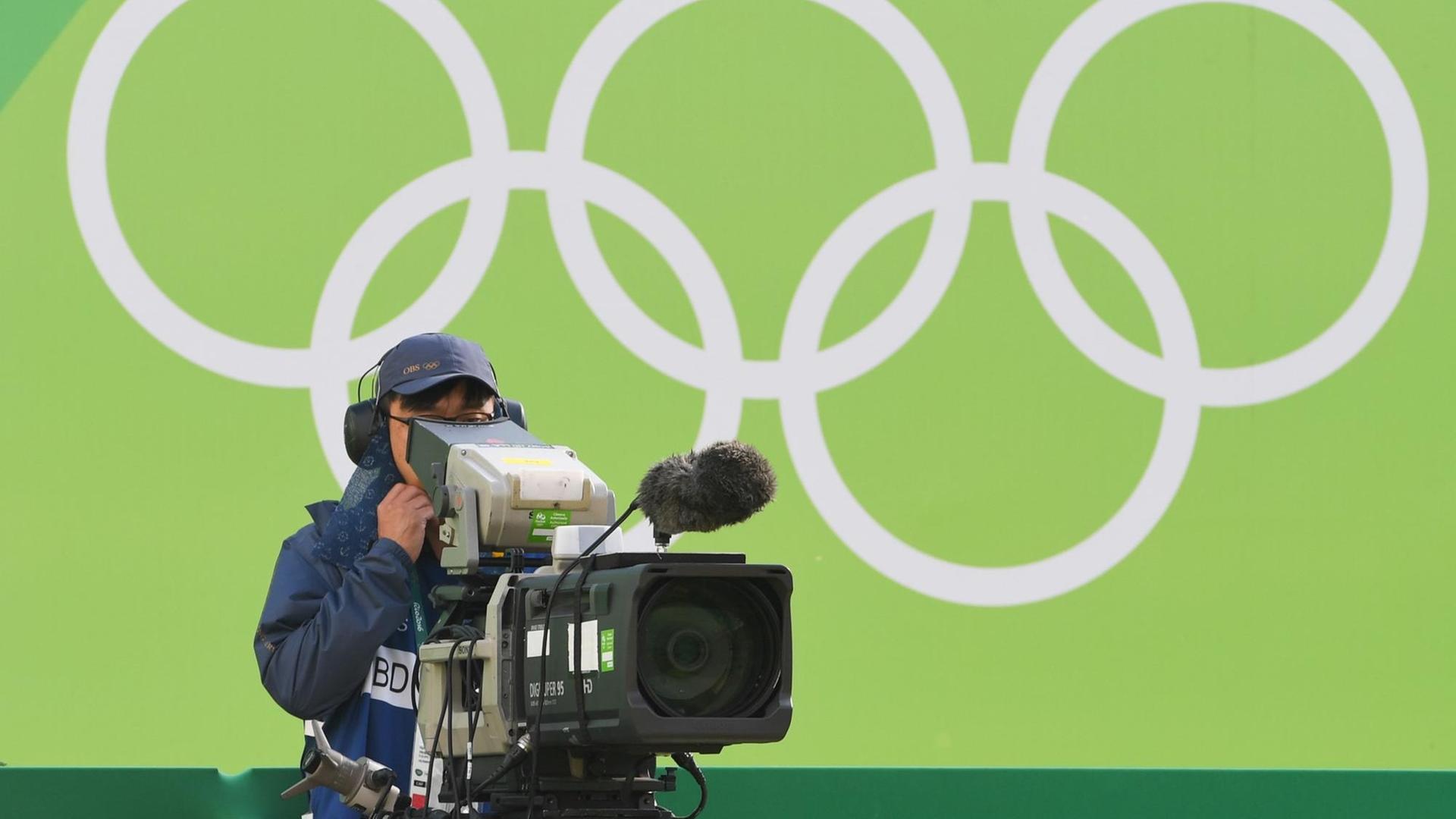 Ein Kameramann steht bei den Olympischen Spielen in Rio vor einer grünen Wand mit den Olympischen Ringen Ein Kameramann steht bei den Olympischen Spielen in Rio vor einer grünen Wand mit den Olympischen Ringen