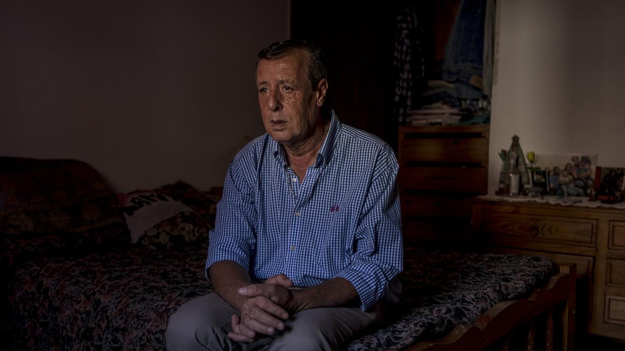 Ein älterer Mann mit blau kariertem Hemd sitzt auf seinem Bett, die Hände zusammengelegt und schaut aus dem Fenster. Ein älterer Mann mit blau kariertem Hemd sitzt auf seinem Bett, die Hände zusammengelegt und schaut aus dem Fenster.