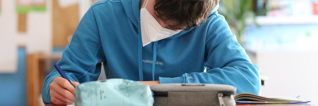 Ein Schüler sitzt mit einem Mundschutz im Klassenzimmer und schreibt. Ein Schüler sitzt mit einem Mundschutz im Klassenzimmer und schreibt.