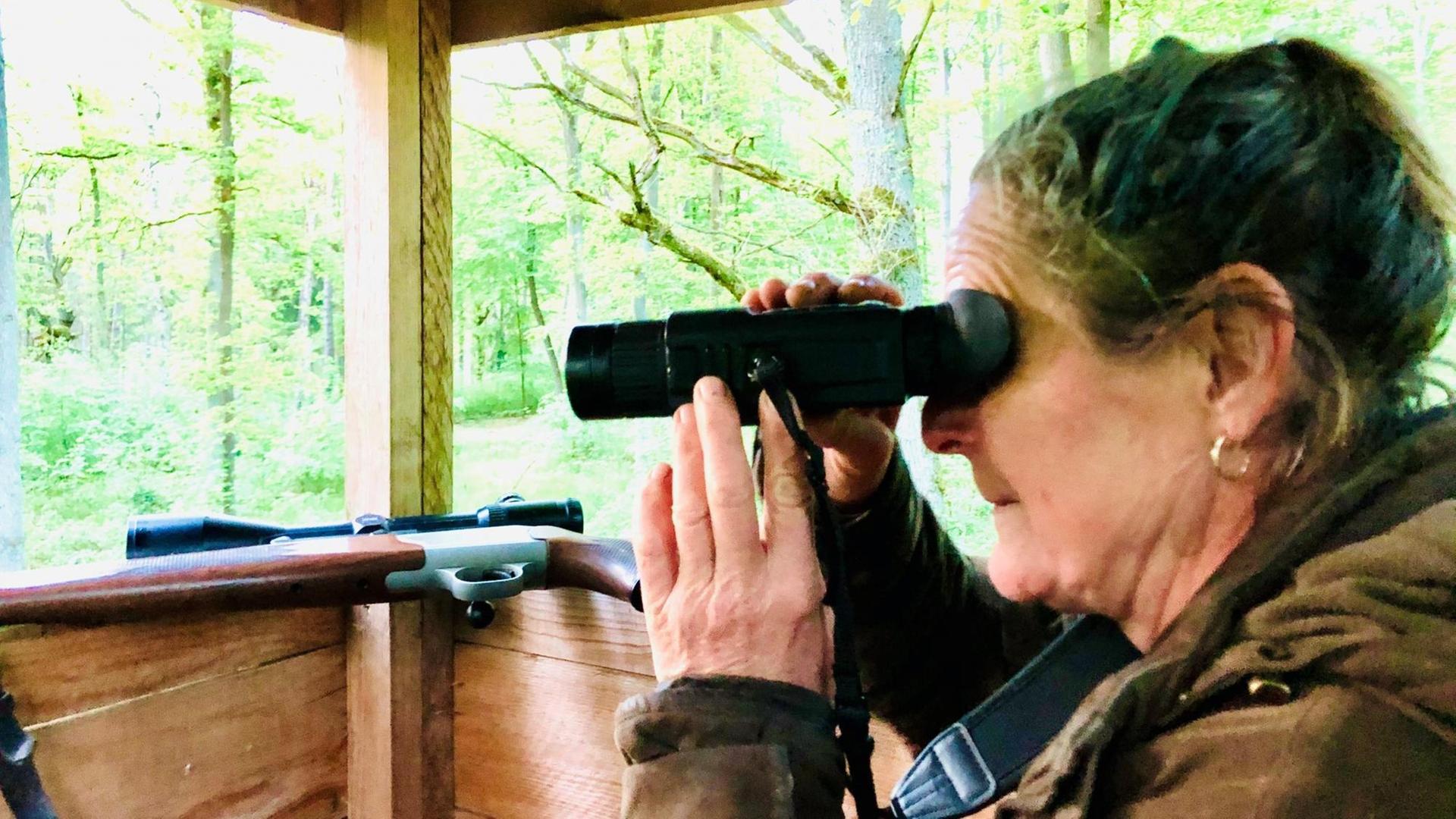 Jägerin Sabine Zuckmantel schaut auf dem Ansitz durchds Fernglas. Im Hintergrund ist ihr Gewehr zu sehen.