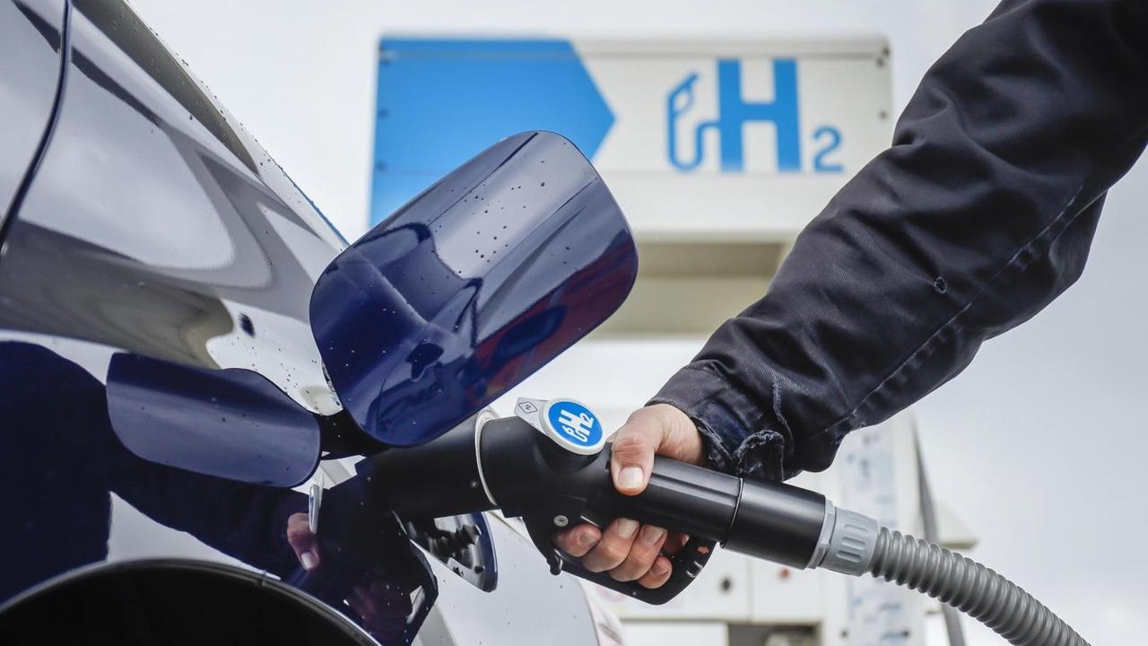 Eine Hand führt den Tankstutzen in ein blaues Auto, im Hintergrund steht eine H2-Tanksäule Eine Hand führt den Tankstutzen in ein blaues Auto, im Hintergrund steht eine H2-Tanksäule