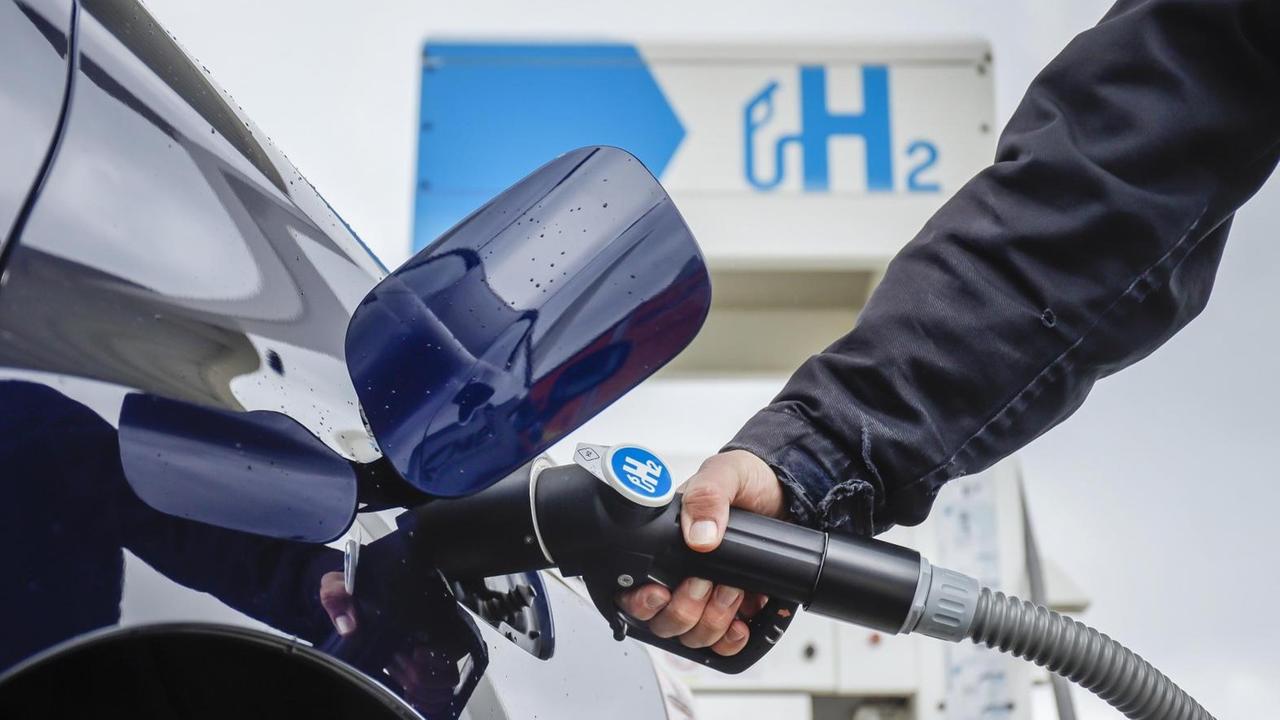 Eine Hand führt den Tankstutzen in ein blaues Auto, im Hintergrund steht eine H2-Tanksäule