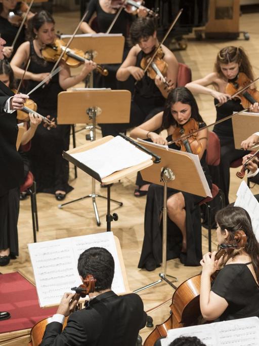 Ein von jungen Frauen dominiertes Orchester sitzt um den Dirigenten, der auf einem roten Pult steht.