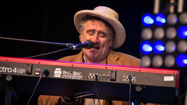Ein Mann sitzt auf einer Bühne am Keyboard, spielt und singt in ein Mikrofon vor seinem Mund.