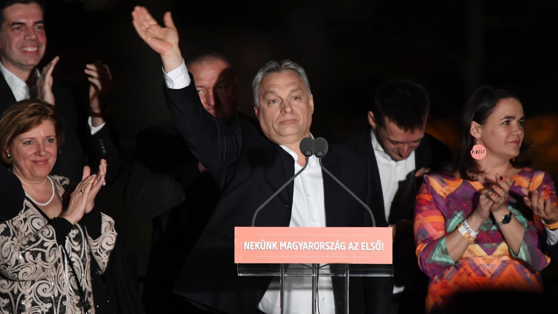 Orban steht im Dunkeln in Freien hinter zwei Mikrofonen und winkte mit erhobener Hand. Daneben zwei Frauen und drei Männer.